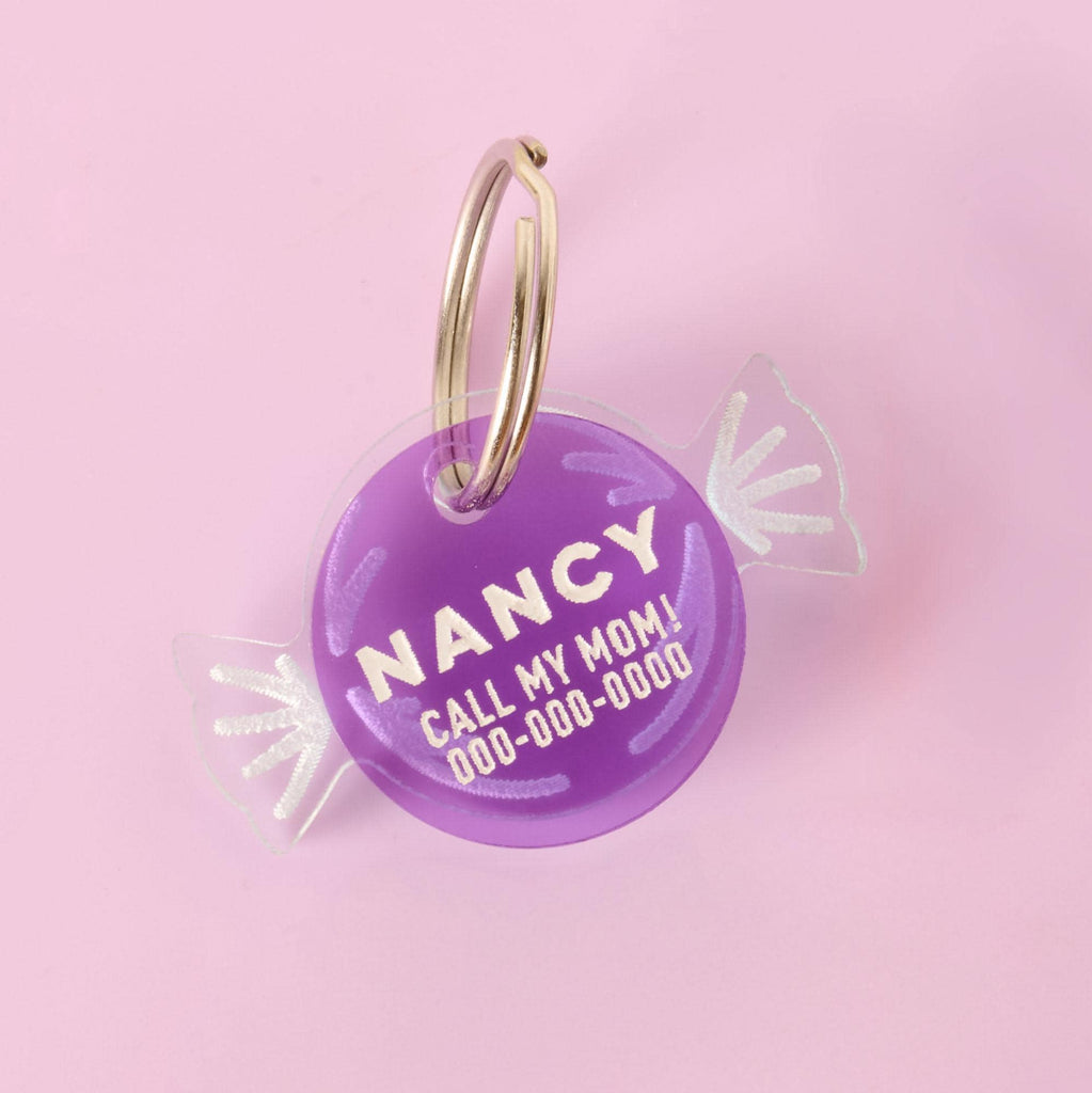 Candy Wrapper Pet Tag Add-on A personalized purple pet ID tag shaped like a wrapped piece of candy. The tag has the name "NANCY" and a placeholder phone number on it. It's attached to a silver ring, perfect for a collar, and is displayed on a solid pink background.
