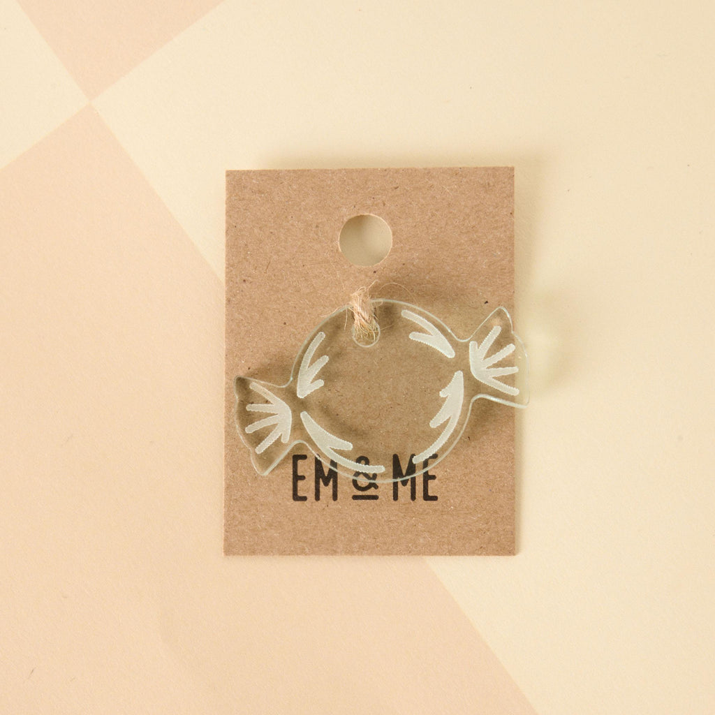 A close-up of a clear, wrapped candy-shaped charm or pin with white design details. The charm is attached to a brown kraft paper card that reads "EM & ME." The item is displayed on a light beige background with a faint checkered pattern.
