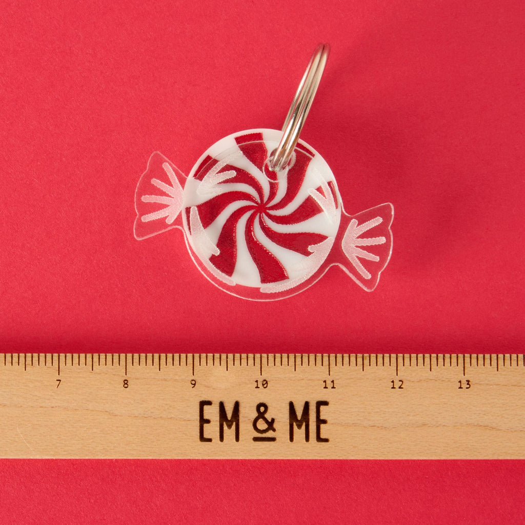 A red and white peppermint swirl candy-shaped charm or keychain in a clear wrapper design. A wooden ruler with the "EM & ME" logo is placed below the charm to show its size. The item is attached to a silver ring and displayed on a solid red background.