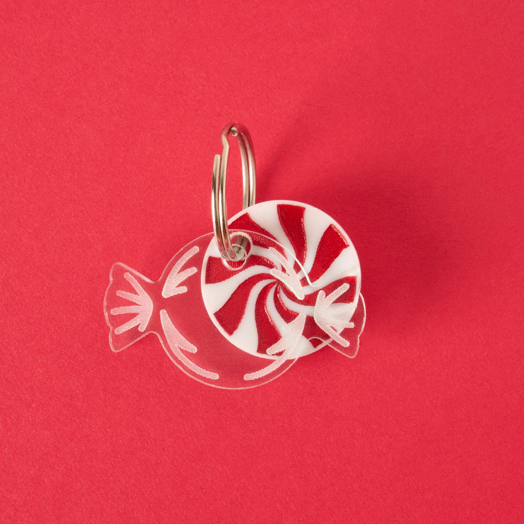 Candy Wrapper Pet Tag Add-on A red and white peppermint candy charm or keychain in a clear wrapper design, attached to a silver ring. The item is displayed on a solid red background.