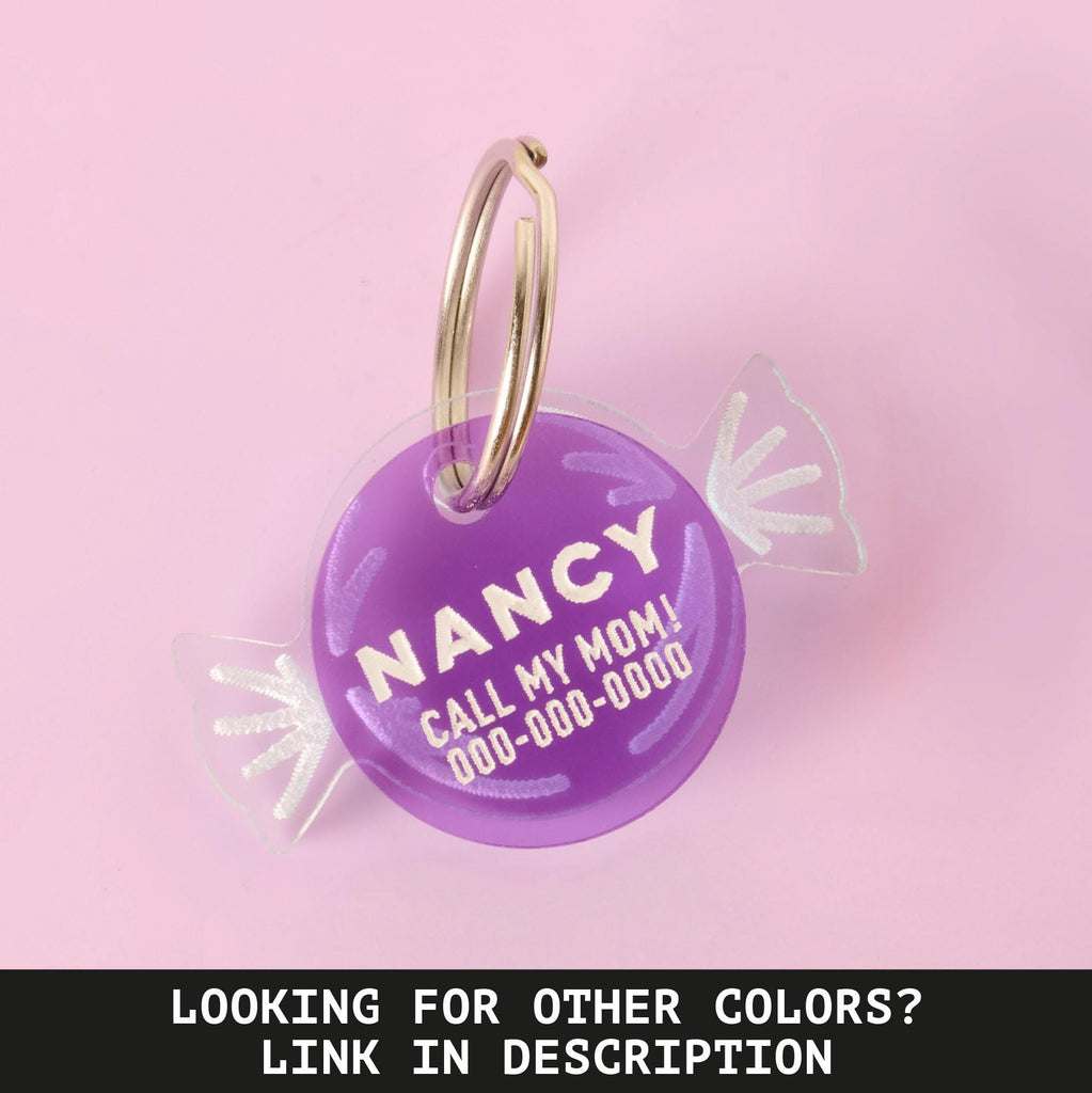 A personalized purple pet ID tag shaped like a wrapped piece of candy. The tag has the name "NANCY" and a placeholder phone number on it. It's attached to a silver ring, perfect for a collar, and is displayed on a solid pink background. A caption at the bottom of the image reads: "LOOKING FOR OTHER COLORS? LINK IN DESCRIPTION".