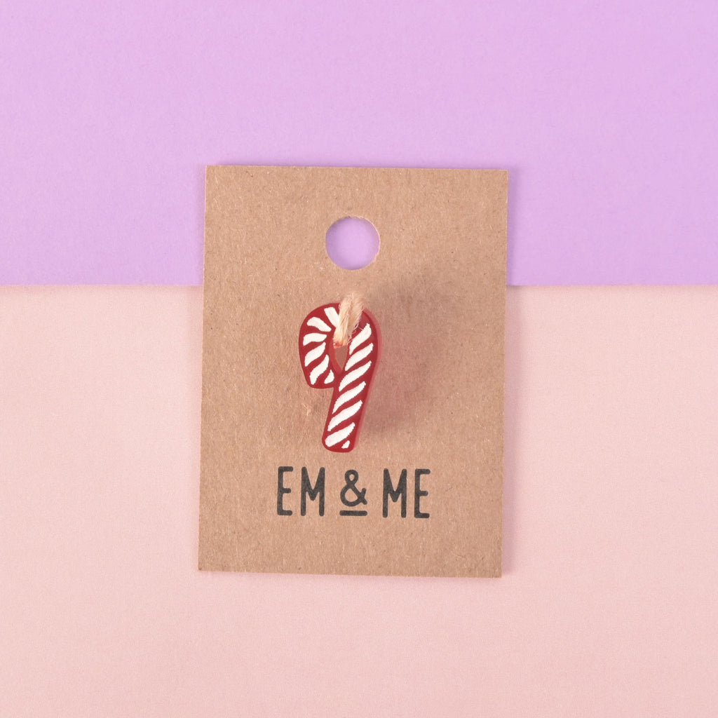 A red and white candy cane-shaped charm or pin attached to a brown paper card that reads "EM & ME." The item is displayed on a background that is half solid purple and half solid pink.