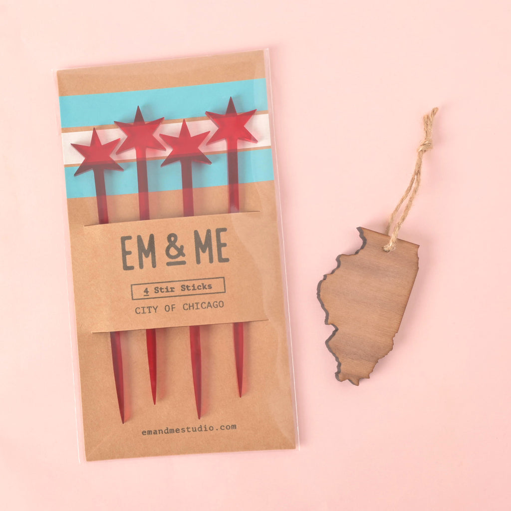 A set of four red cocktail stirrers with Chicago Flag stars on top, packaged with a brown card that features the Chicago Flag design and the text "CITY OF CHICAGO." Beside the package is a wooden ornament or keychain shaped like the state of Illinois. Both items are from "EM & ME" and are displayed on a solid pink background.