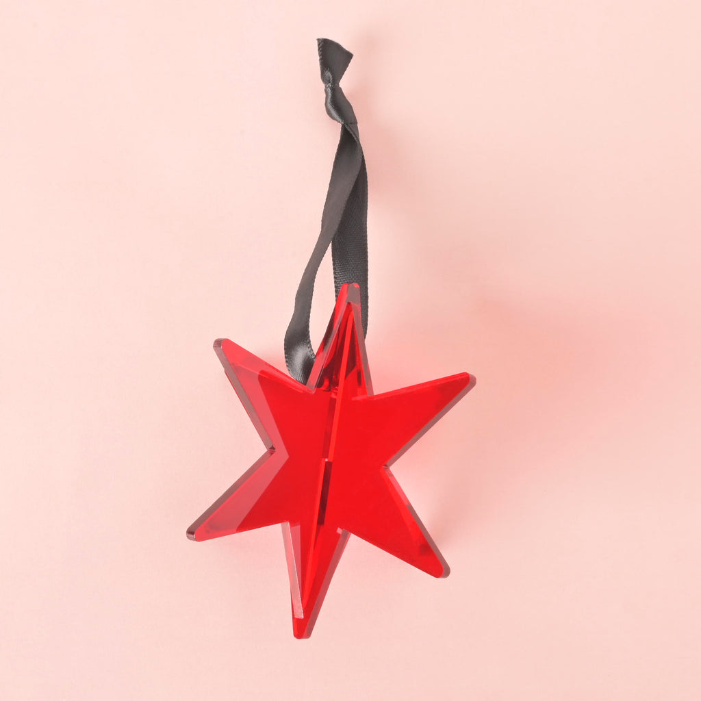 Red acrylic Chicago star ornament with a black ribbon hanger, displayed on a pastel pink background, perfect for holiday decor.