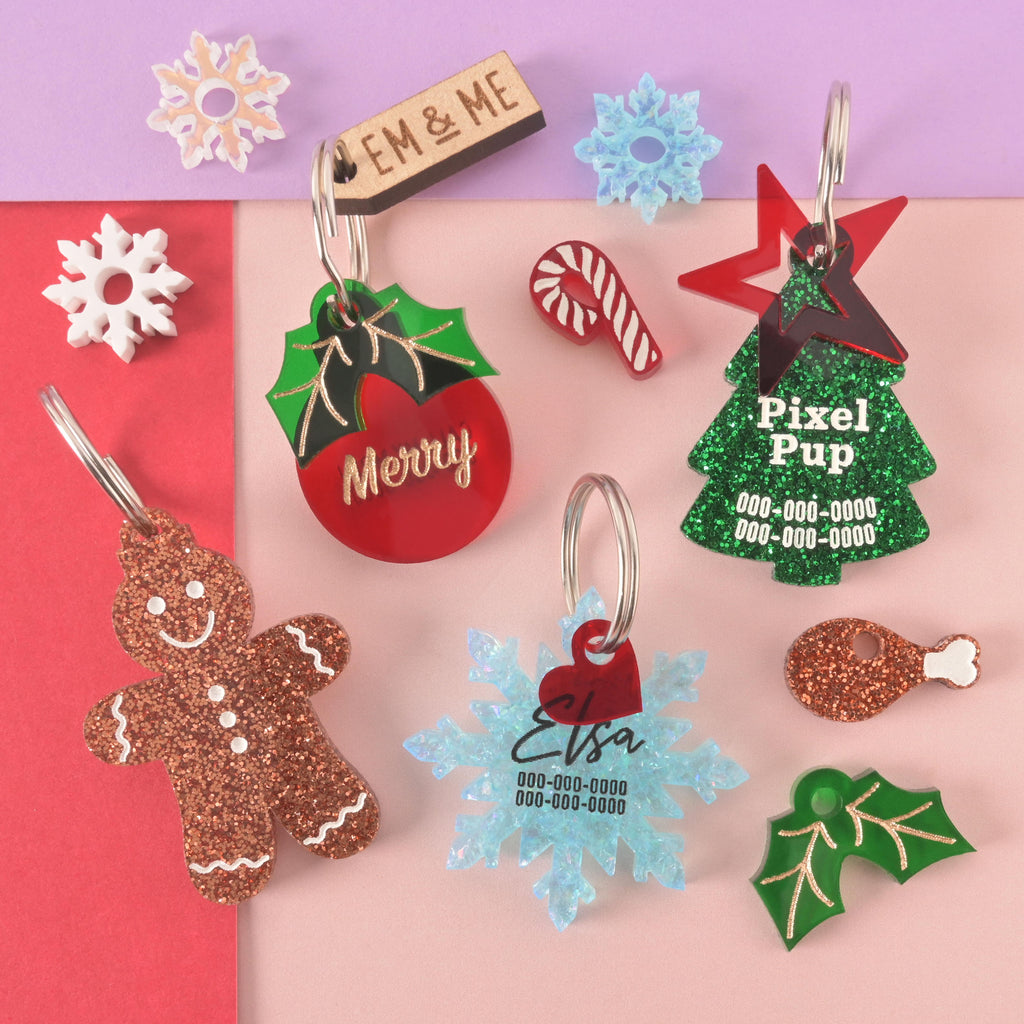 A collection of assorted holiday-themed pet tags and charms scattered on a pink, red, and purple background. The items include a glittery copper gingerbread man, a red ornament tag that says "Merry," a green glitter Christmas tree tag for "Pixel Pup," a blue glitter snowflake tag for "Elsa," and a wooden charm that says "EM & ME." Surrounding them are small charms like snowflakes, a candy cane, holly leaves, and a drumstick.