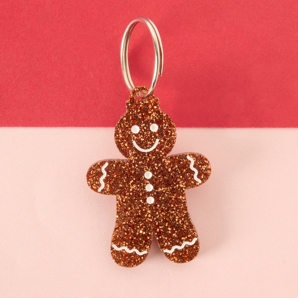 Close-up of a gingerbread man-shaped acrylic pet ID tag, featuring a brown glitter finish and white frosting details. The tag is attached to a silver split ring and displayed on a color-blocked background of light pink and bright red.