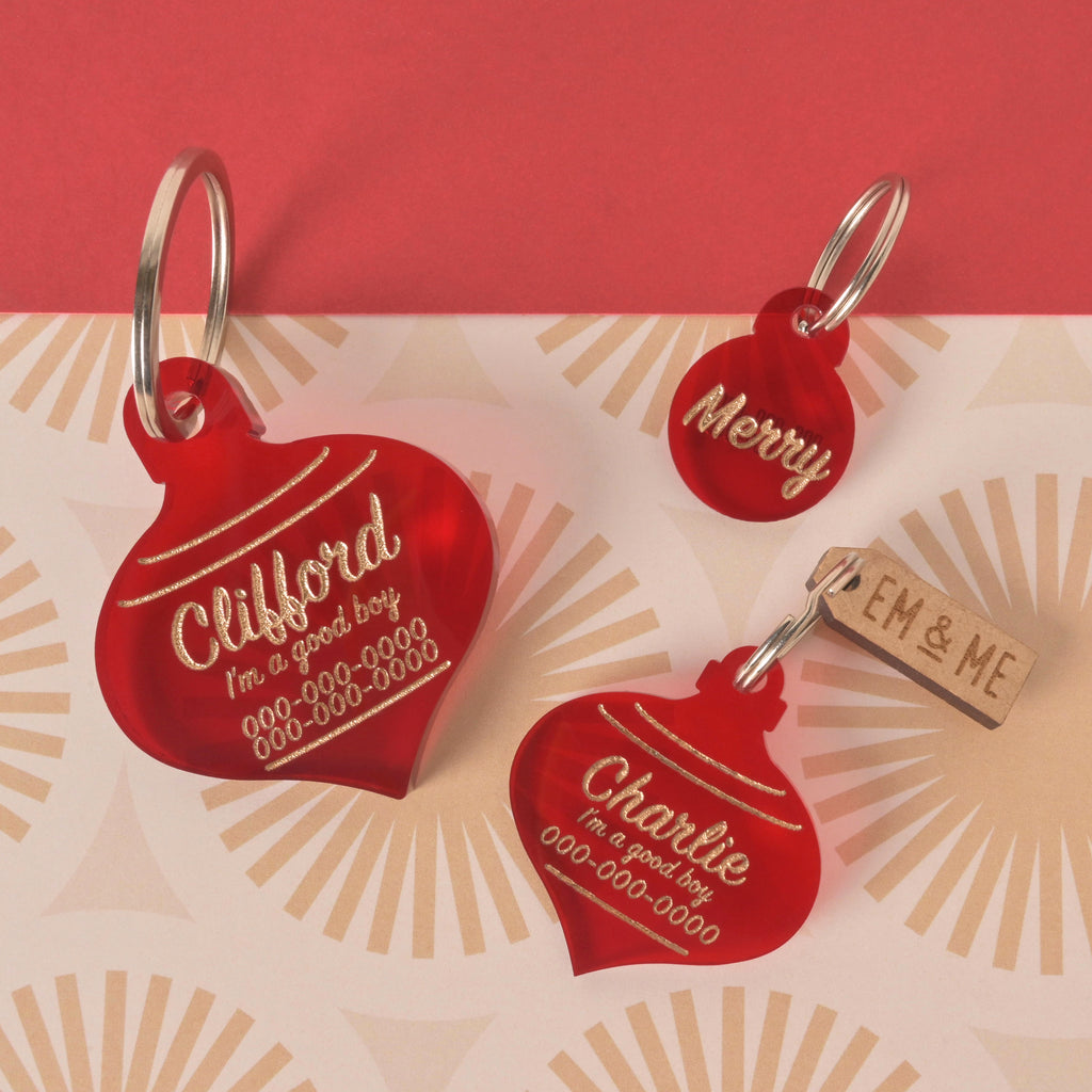 A trio of custom, red, Christmas ornament-shaped pet ID tags. The two larger tags are engraved with "Clifford" and "Charlie" along with "I'm a good boy" and a phone number. The smallest tag says "Merry." A small wooden tag with "EM & ME" is also visible. The tags are arranged on a red and gold patterned background.