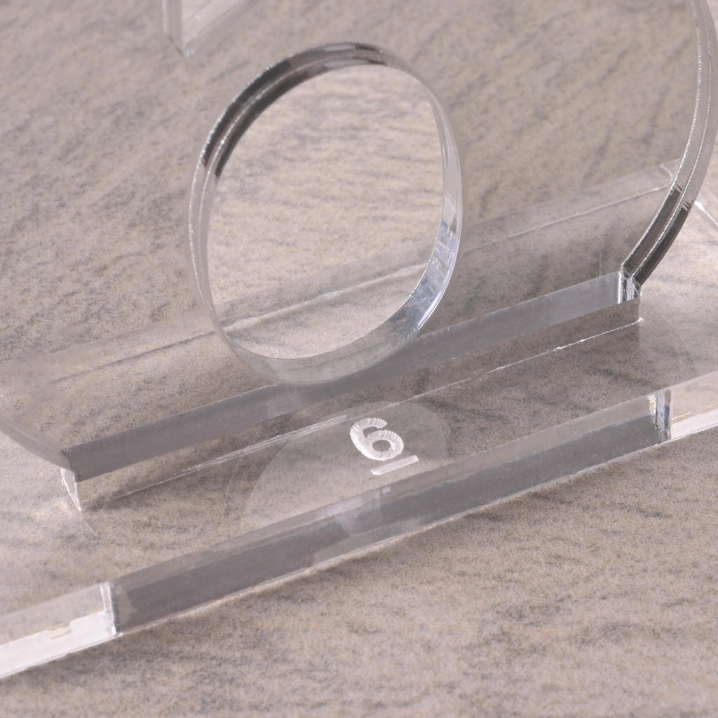 Close-up of a modern clear acrylic table number “6” with a rectangular acrylic base on a neutral stone-like surface.