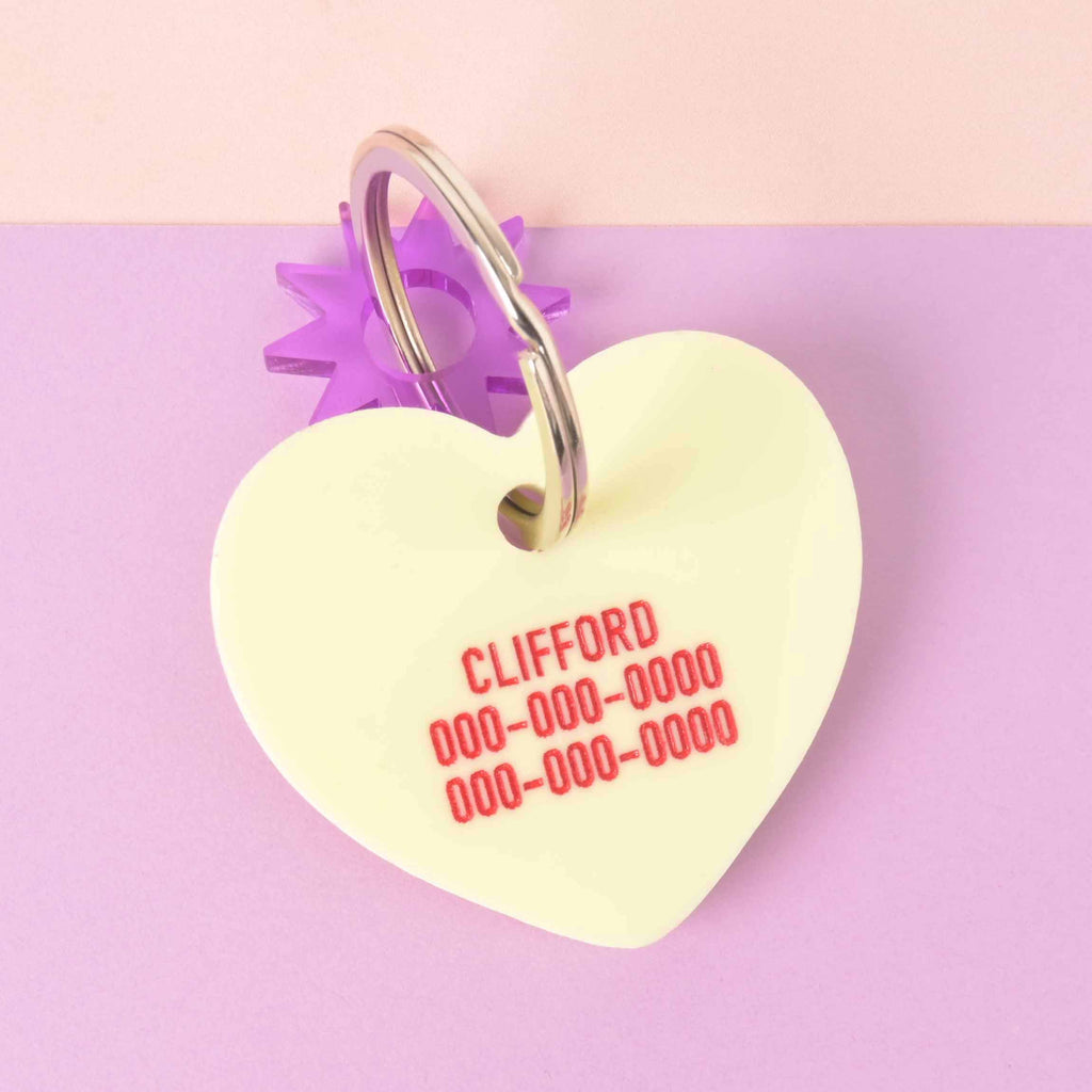 Clifford pastel yellow heart-shaped pet ID tag with custom red engraved contact details, silver ring, and purple star connector on lavender-pink.