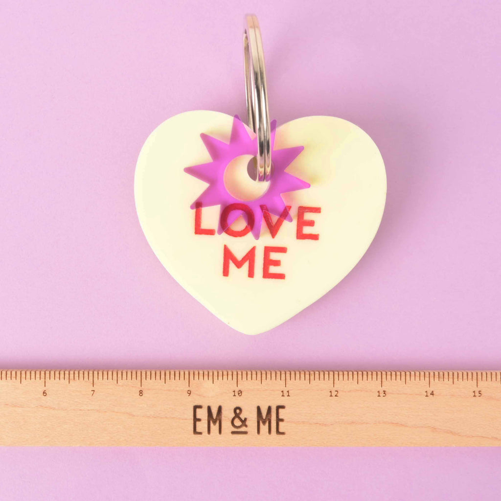 Clifford pastel yellow heart-shaped pet ID tag with red “LOVE ME” engraving, purple star connector, silver ring, and ruler for size scale.