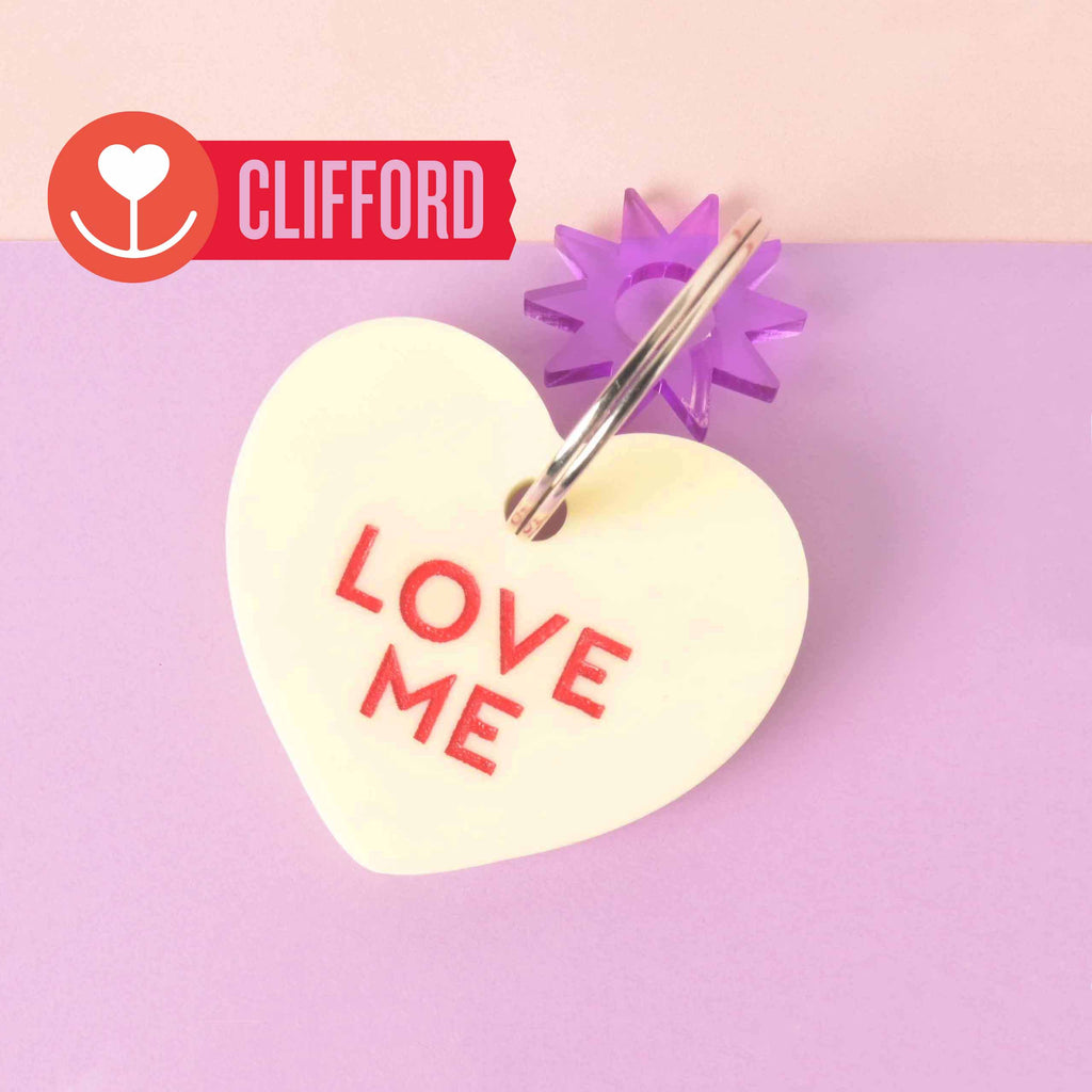 Clifford pastel yellow heart-shaped pet tag with bold red “Love Me” text, silver ring, and purple star connector on a lavender-pink backdrop.