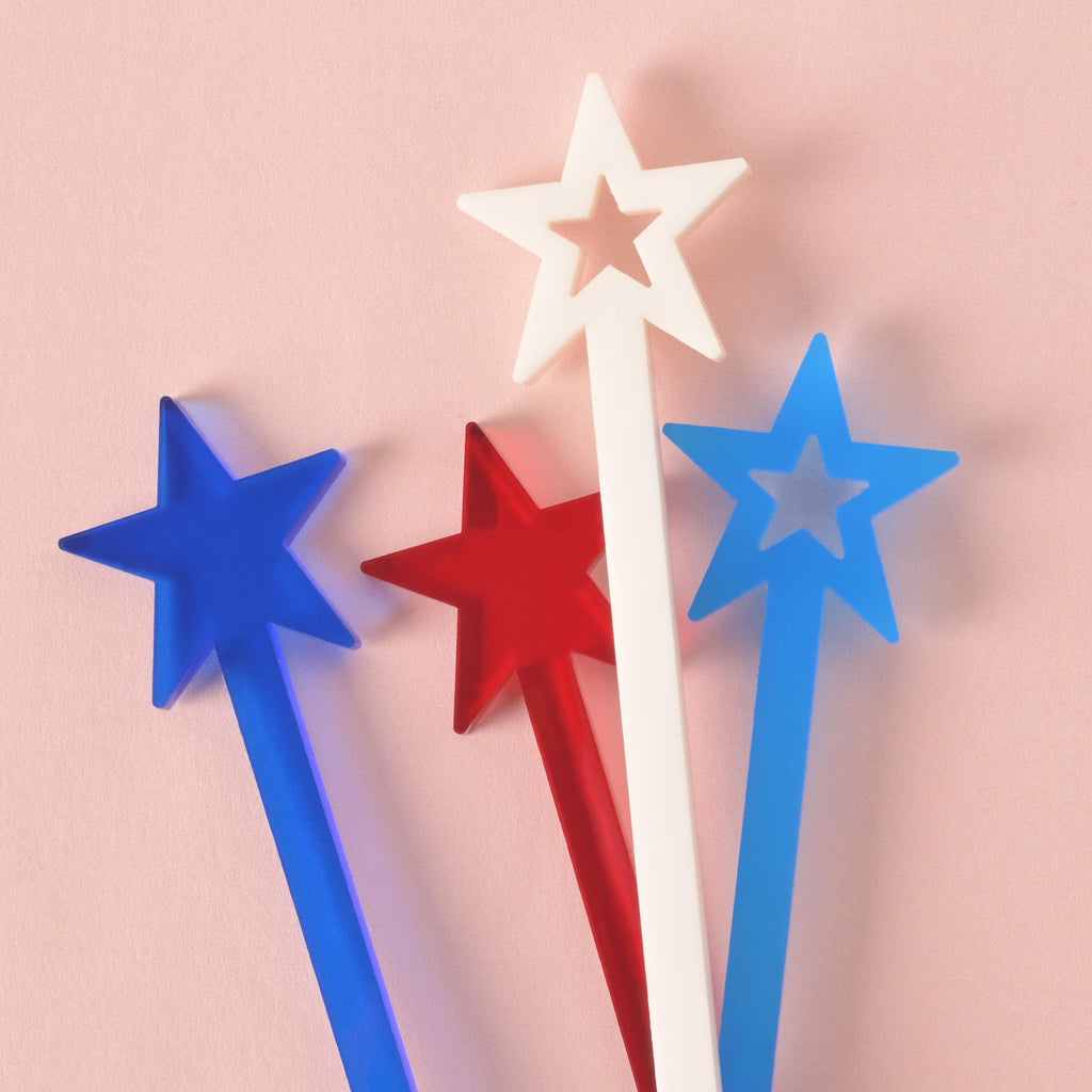 A collection of four red, white, and blue star-shaped cocktail stirrers. The stirrers are positioned in a scattered group against a solid pink background.