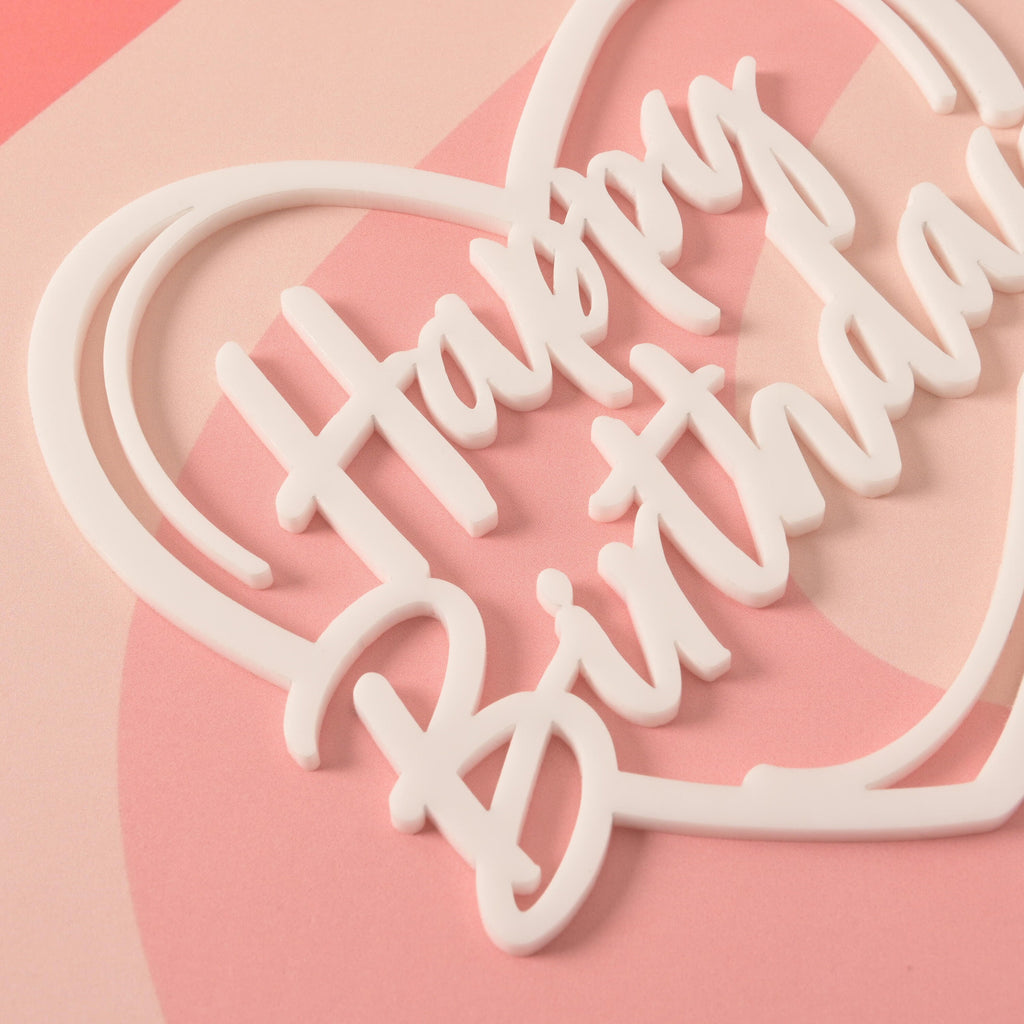 A close-up of a white cake topper with the words "Happy Birthday" written in a curvy font. The topper is on a pink and white striped background.