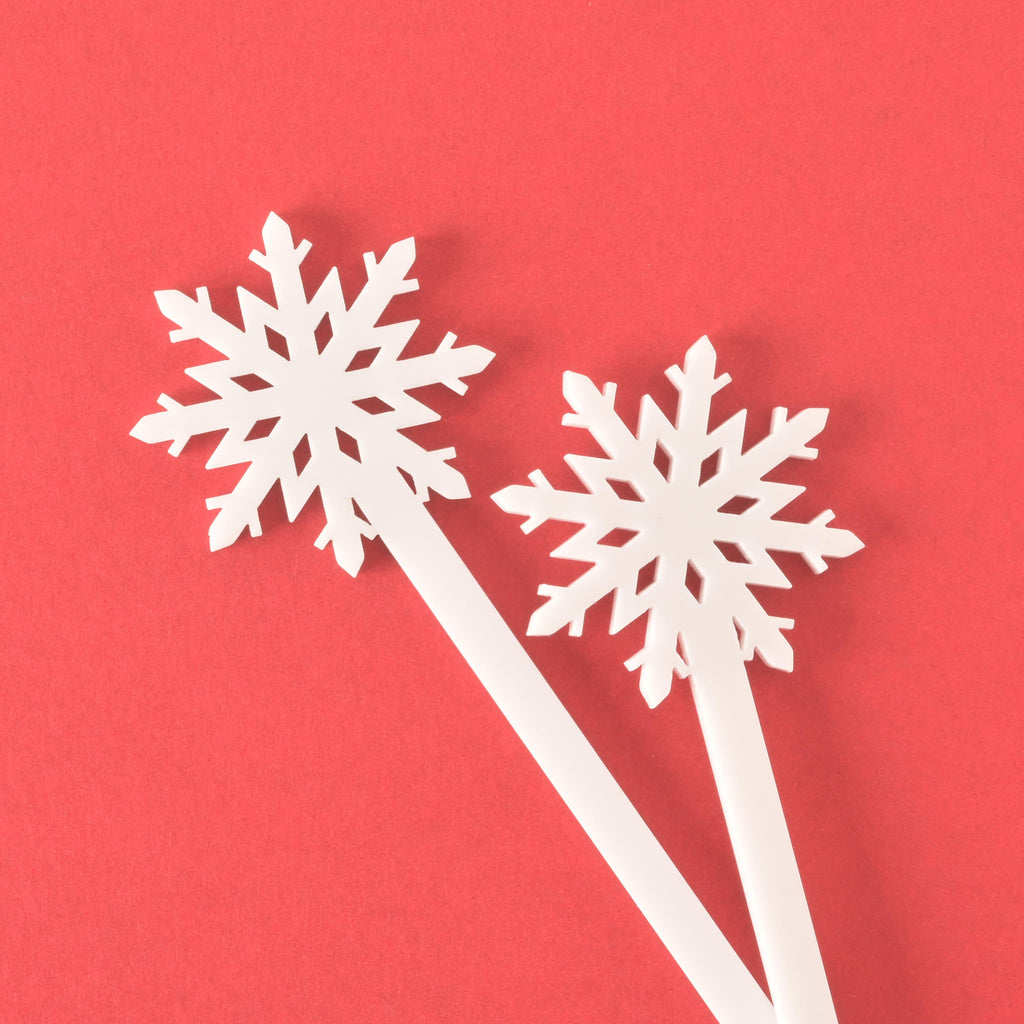 A close-up of two white snowflake-topped cocktail stirrers, overlapping on a solid red background. The stirrers have a clean, solid white appearance.