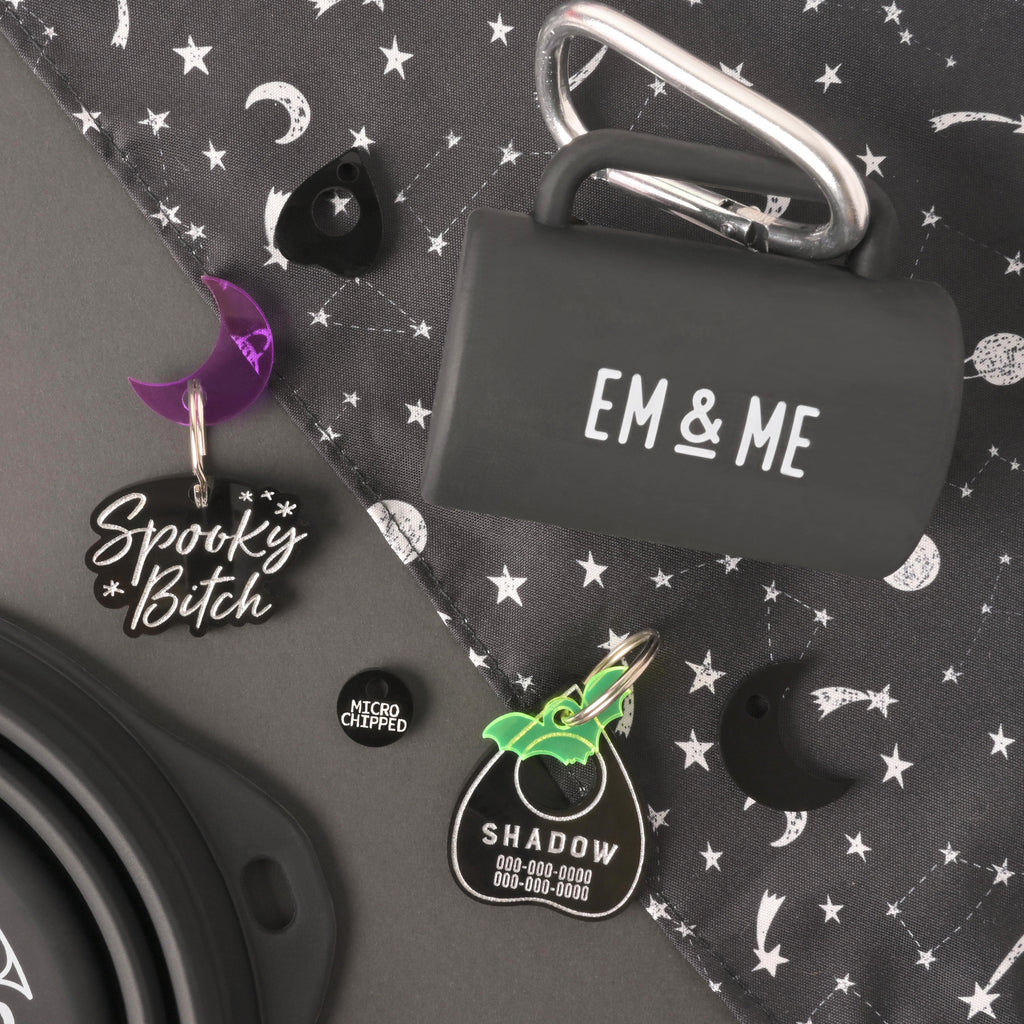 Collection of black pert products. Poop bag holder that reads "EM & ME", a personalized pet tag that says "spooky bitch" on the front with a purple moon charm, ouija planchet-shaped pet tag with a fluorescent green bat mini and a "microchipped" mini on a black background with silver stars and planets