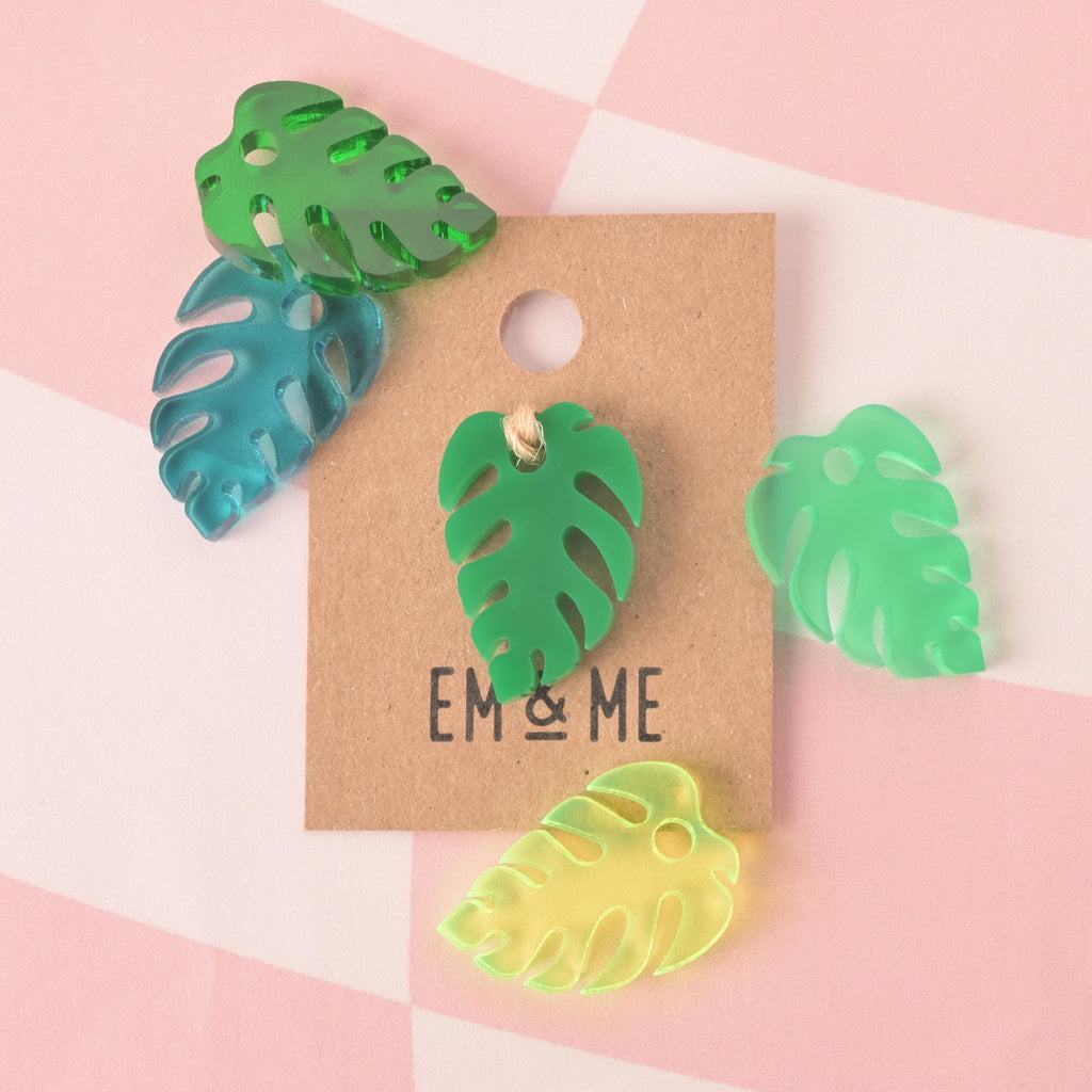 A collection of five Monstera leaf-shaped charms or pins in assorted translucent green and blue colors. One dark green charm is attached to a brown kraft paper card that reads "EM & ME." The items are scattered on a pink and white checkered background.