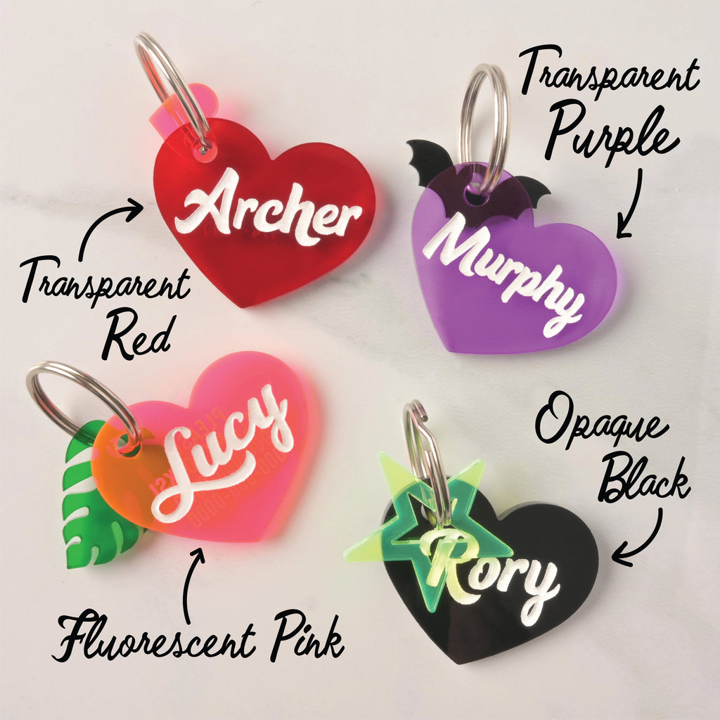 Four custom plastic heart-shaped pet ID tags labeled with pet names Archer, Murphy, Lucy, and Rory, shown in color options: Transparent Red, Fluorescent Pink, Transparent Purple, and Opaque Black, each with themed accent charms