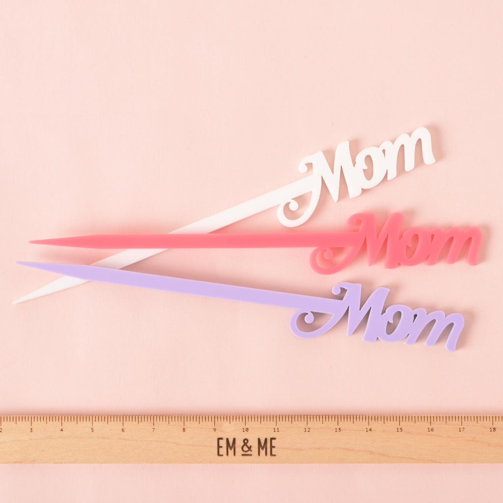 three "Mom" cocktail stirrers in different colors—white, pink, and lavender—are shown next to a ruler with the "EM & ME" logo to show their size. The stirrers are arranged in a fanned pattern against a solid pink background.