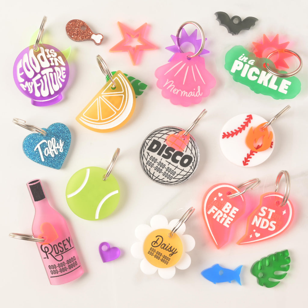 Assorted acrylic pet ID tags in playful designs including wine bottle, lemon slice, tennis ball, disco ball, daisy, baseball, best friends hearts, pickle, glitter heart, fish, bat, leaf, and more, each engraved with pet name and phone number.