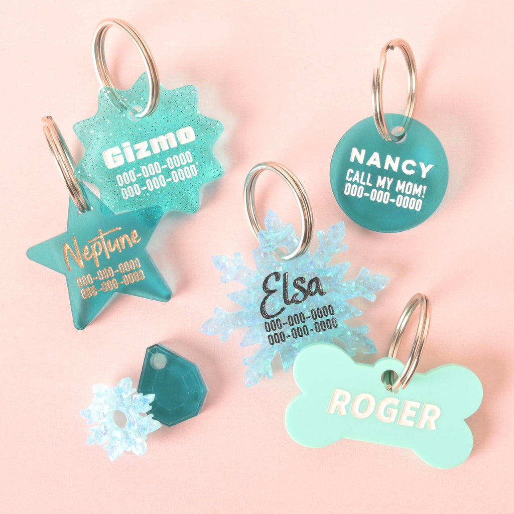 Assorted custom Teal acrylic pet ID tags in star, snowflake, circle, hexagon, and bone shapes with engraved names and phone numbers, displayed on a pink background.