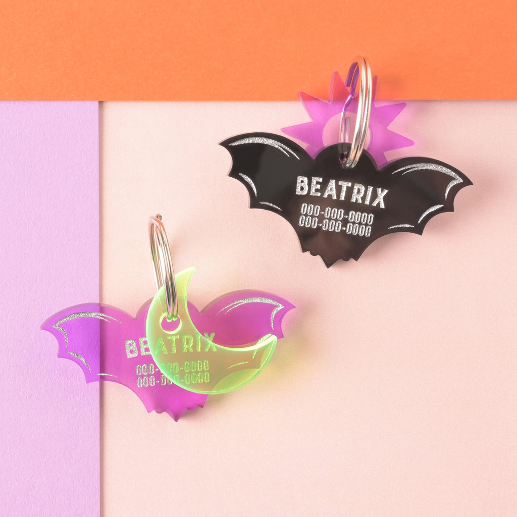 Two personalized bat-shaped pet ID tags for 'BEATRIX' shown together: one is opaque black acrylic with a translucent purple sunburst accent charm, and the other is translucent purple acrylic layered with a neon yellow crescent moon charm.