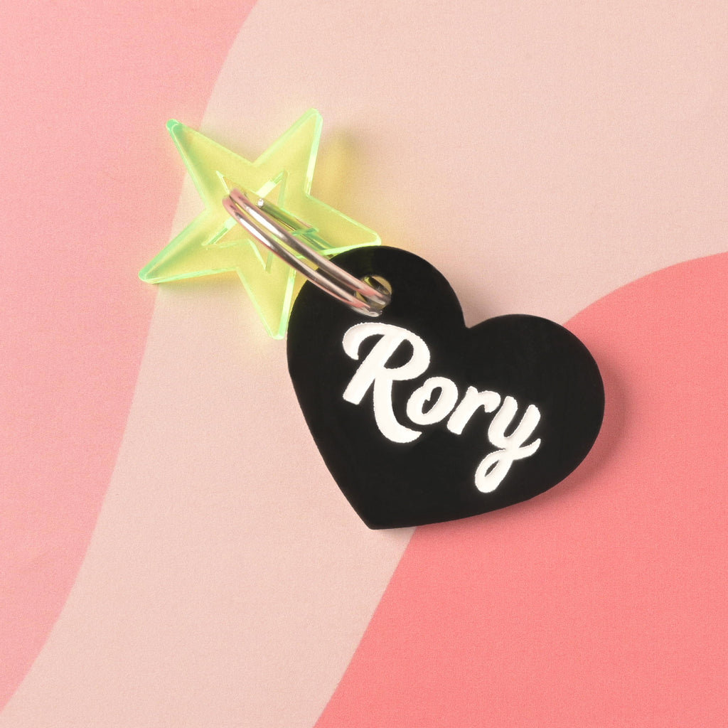 Black acrylic heart-shaped pet ID tag with the name Rory in white script, paired with a neon green star charm on a pink abstract background