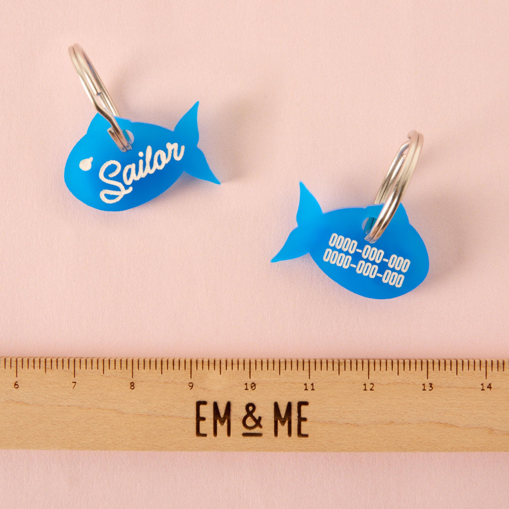 Front and back of blue fish-shaped pet ID tags, one side with “Sailor” name and the other engraved with phone numbers, shown next to a ruler for size reference.