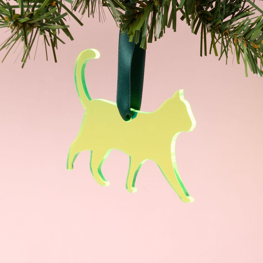A laser-cut fluorescent neon yellow-green acrylic cat silhouette ornament, hanging by a dark green ribbon from a pine branch, set against a pink background.