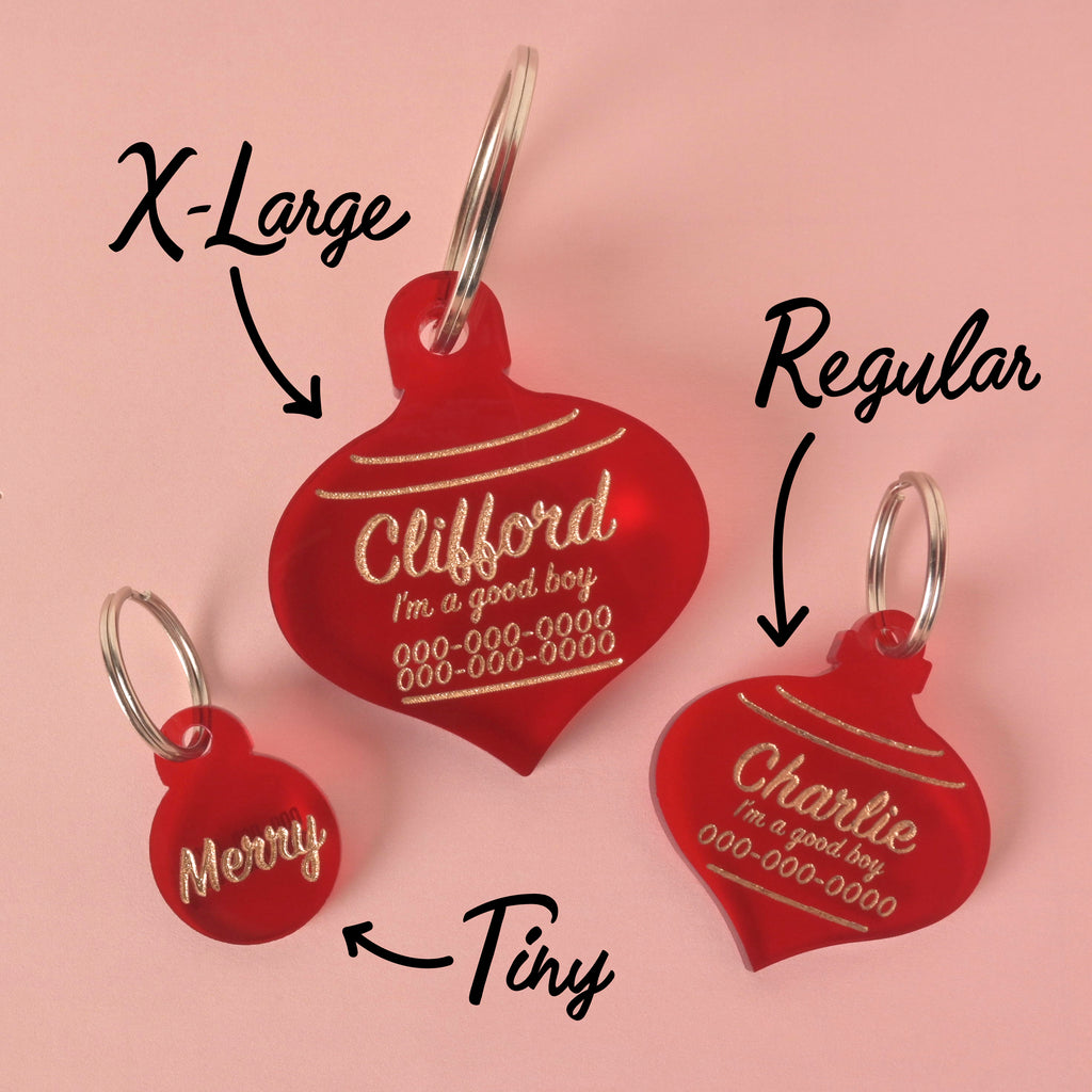 A promotional image showing three red, Christmas ornament-shaped pet ID tags of varying sizes: Tiny, Regular, and X-Large. The tags are engraved with different information, including the names "Merry," "Charlie," and "Clifford," along with the phrase "I'm a good boy" and a phone number. Arrows point to each tag, indicating its size. All tags have silver keyrings and are displayed on a solid pink background