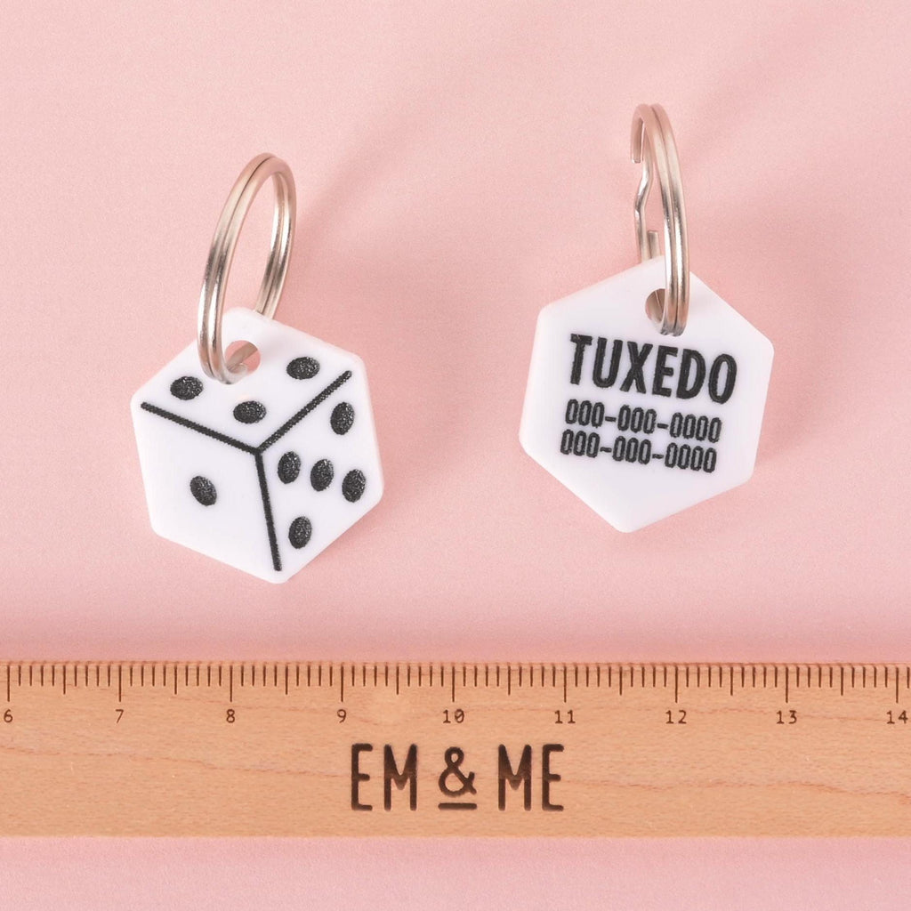 Two dice-shaped pet ID tags with engraving, one showing black dice design and the other customized with pet name Tuxedo and phone number, displayed with ruler for size reference.
