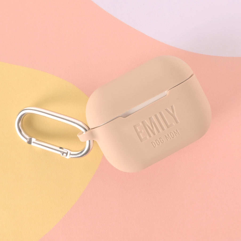 A close-up shot of a beige or tan AirPods case. The case has a shiny silver carabiner clip attached. The front of the case is custom-engraved with "EMILY" in large letters and "DOG MOM" below it. The background is a pastel peach and yellow.