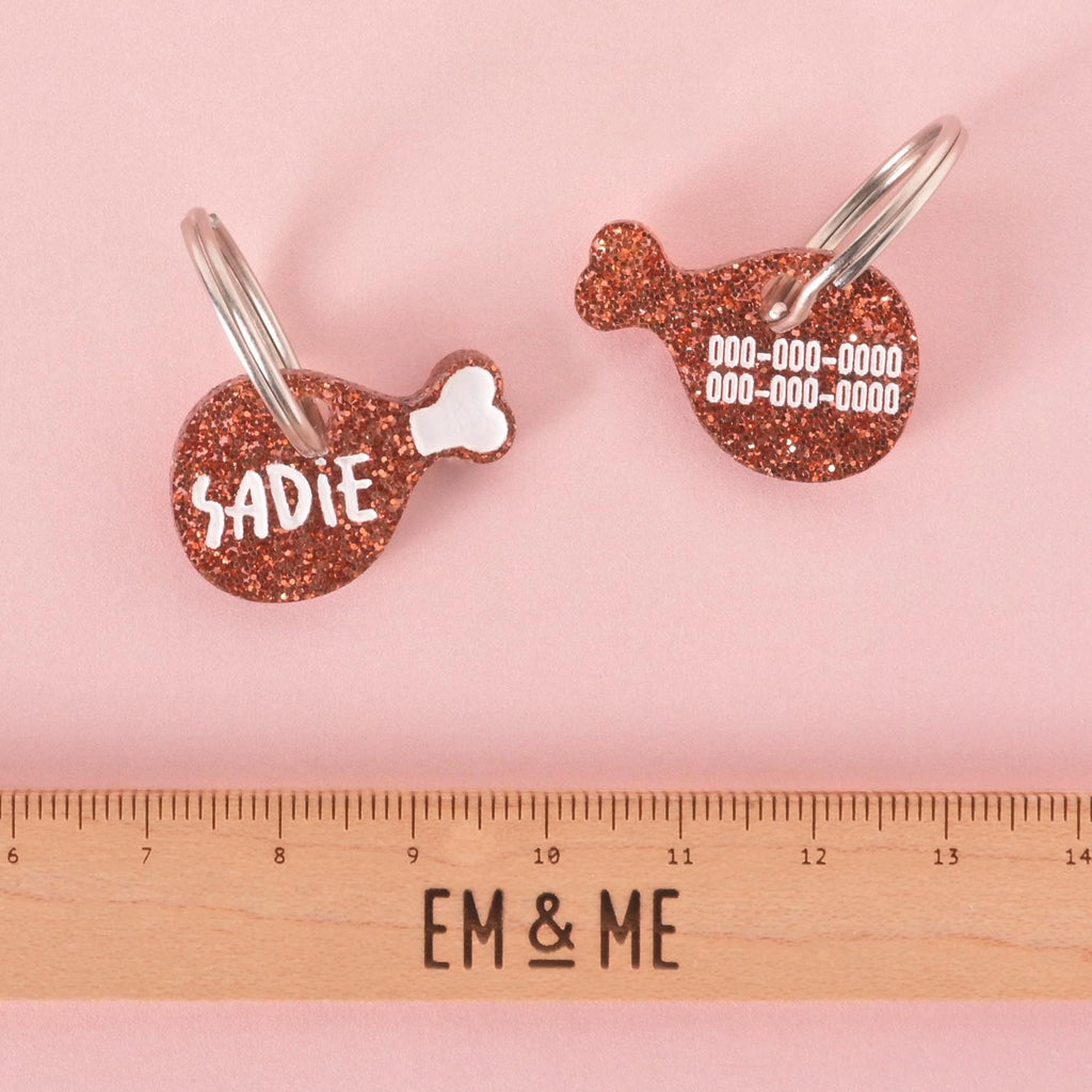 Two glitter drumstick-shaped pet ID tags displayed on a pink background above a wooden EM & ME ruler. One side shows the name “Sadie” and the other side displays placeholder phone numbers.