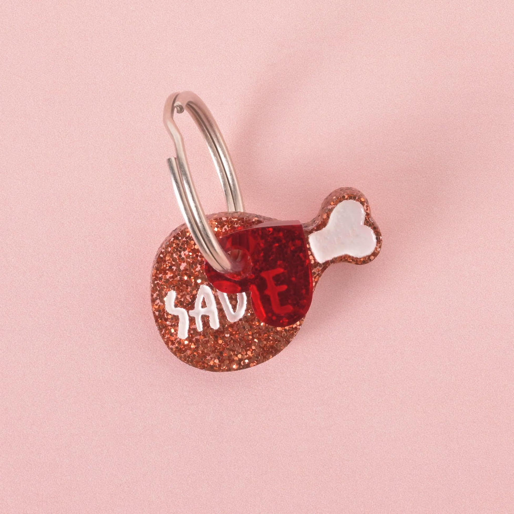 Small glittery drumstick-shaped pet ID tag with the name “Sadie” in white text, attached to a silver ring, displayed on a pink background.
