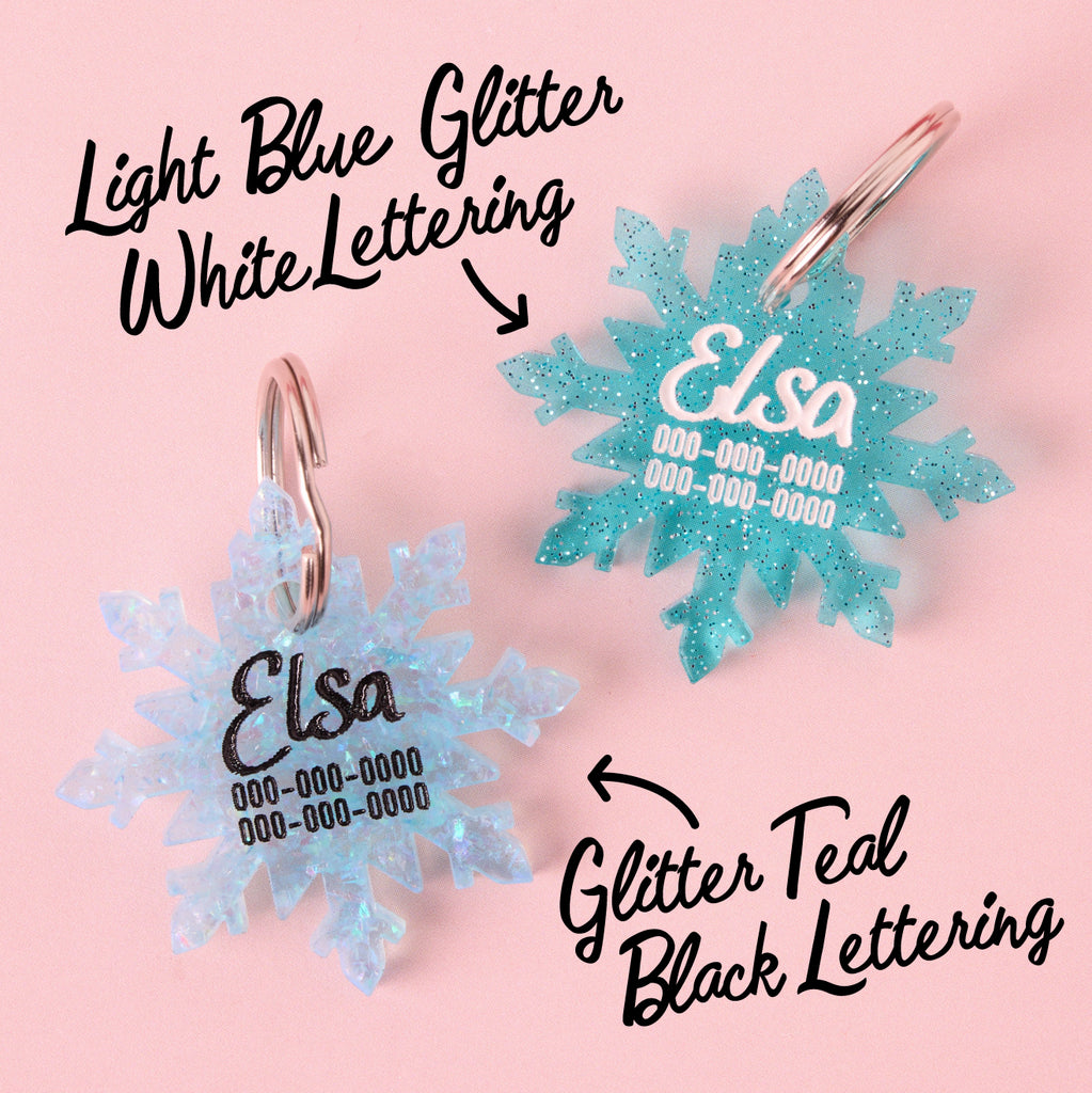 A comparison of two custom snowflake-shaped pet ID tags engraved for 'Elsa'. One is Glitter Light Blue with White Lettering, and the other is Glitter Teal with Black Lettering, demonstrating the available acrylic and engraving options.