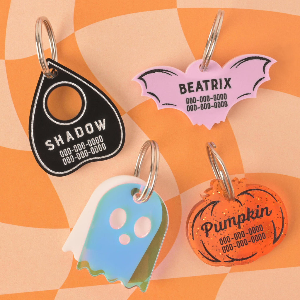 A group of four custom, spooky-themed pet ID tags with silver keyrings. The tags include a black planchette tag engraved with "SHADOW," a pink bat tag with "BEATRIX" and pink details, a translucent blue and white ghost tag, and an orange glitter pumpkin tag engraved with "Pumpkin." All tags are engraved with a phone number and are arranged on an orange and beige checkered background.