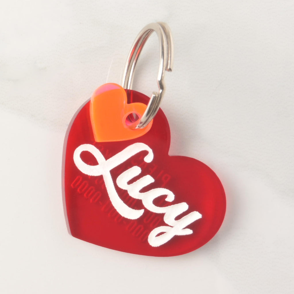 Red acrylic heart-shaped pet ID tag with the name Lucy in white script, featuring a small orange heart accent and metal keyring