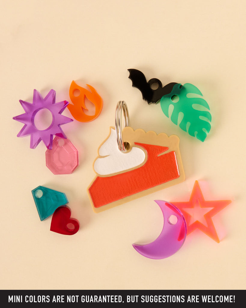 A central, layered pie slice pet tag with whipped cream, surrounded by an assortment of mini acrylic charms: a black bat, tropical leaf, purple sunburst, crescent moon, gemstones, a red heart, and an orange flame. The image highlights the charm customization options.