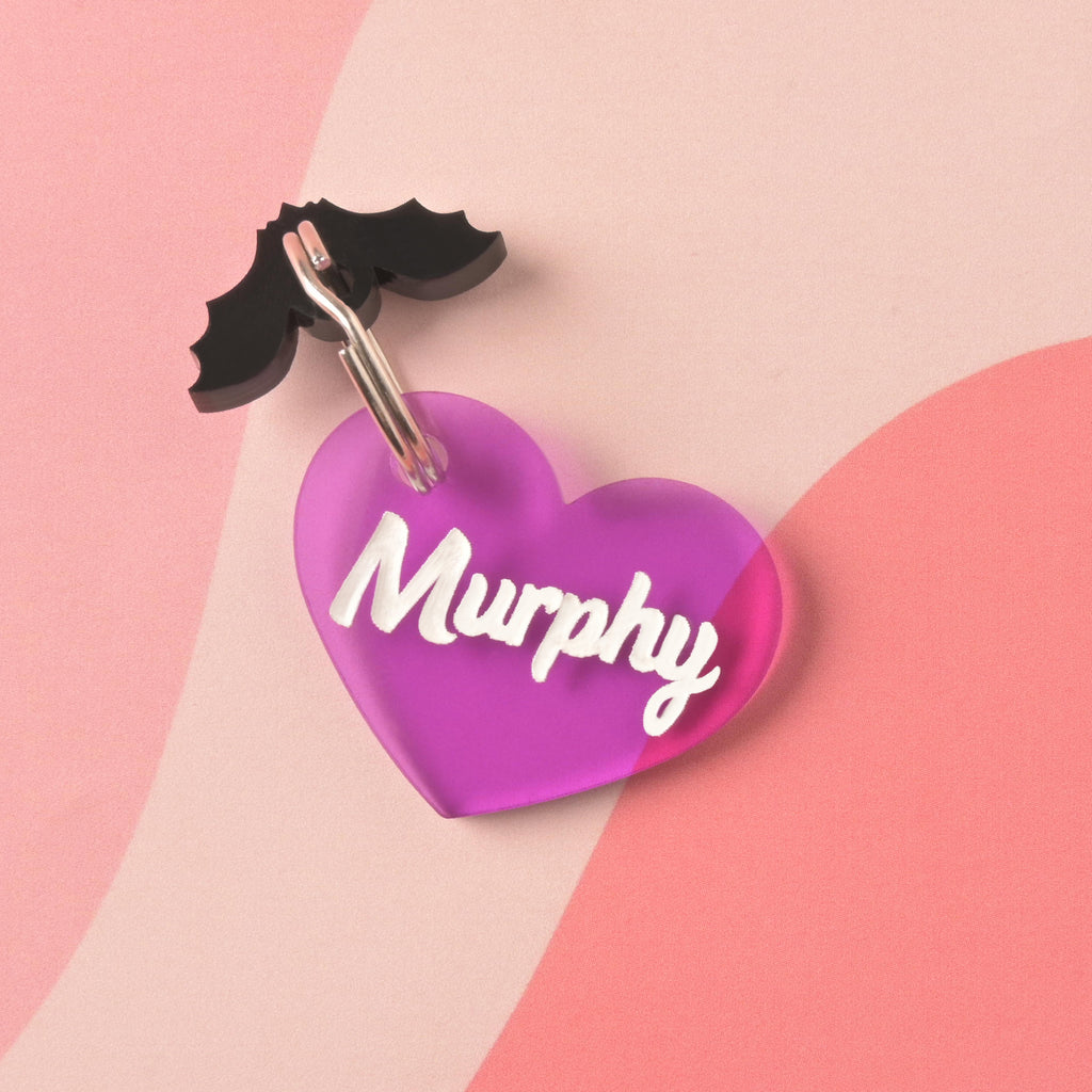 Purple acrylic heart-shaped pet ID tag with the name Murphy in white script, paired with a black bat wing charm, displayed on a pink and coral abstract background