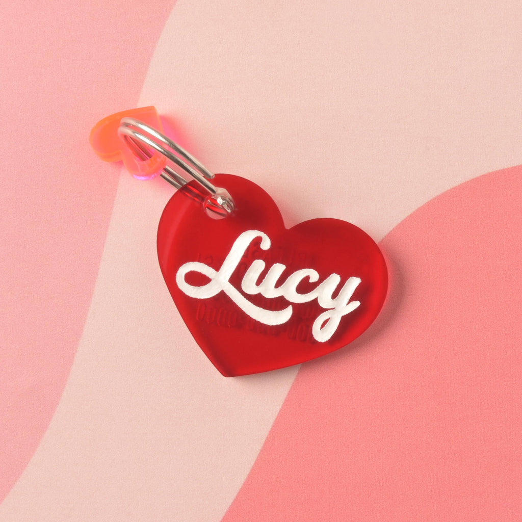 Red acrylic heart-shaped pet ID tag with the name Lucy in white script, attached to a metal ring and orange heart charm, displayed on a pink abstract background