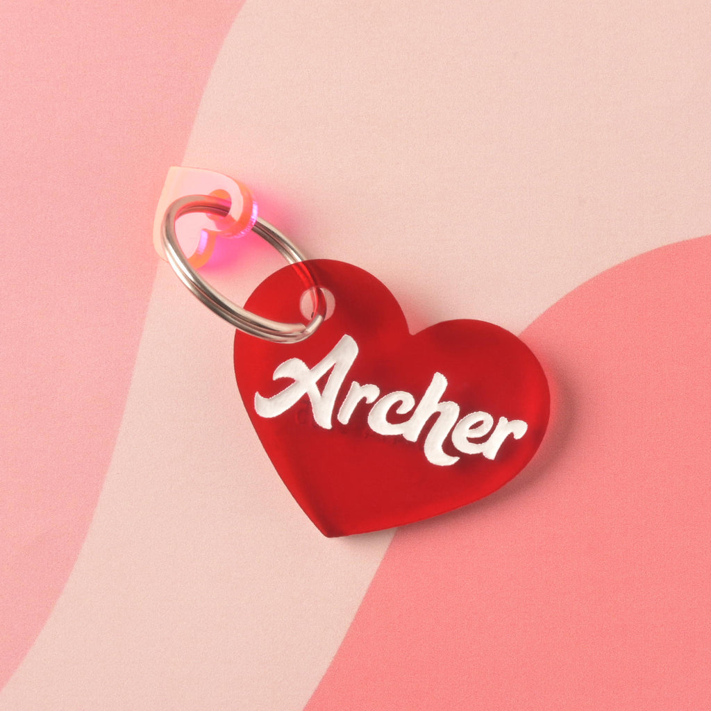Red acrylic heart-shaped pet ID tag with the name Archer in bold white script, paired with a translucent pink heart charm, displayed on a pink abstract background
