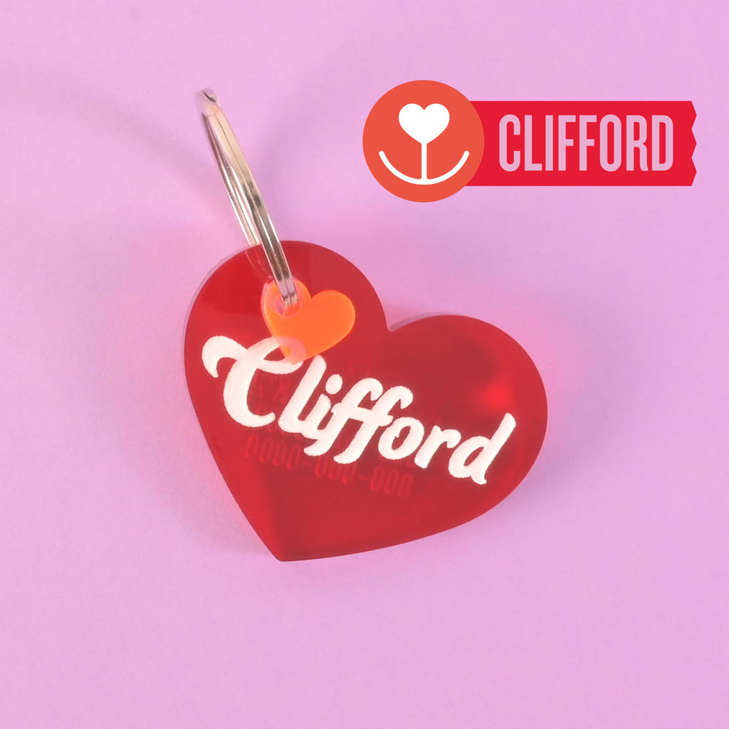 A red heart-shaped pet ID tag with the name 'Clifford' written in a white, hand-drawn script font on the front, and a small red heart-shaped seasonal mini attached to it.