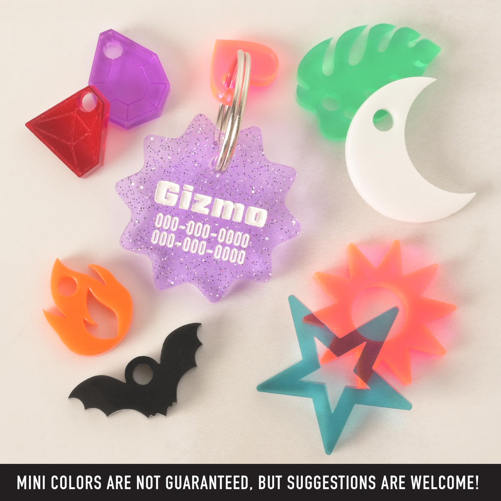 A purple, glittery, starburst-shaped pet ID tag with "Gizmo" and a phone number engraved on it. The tag is surrounded by a variety of smaller charms, including a red gem, a purple gem, an orange flame, a black bat, a turquoise star, an orange star, a green tropical leaf, a white crescent moon, and an orange-pink heart. The items are arranged on a white background.