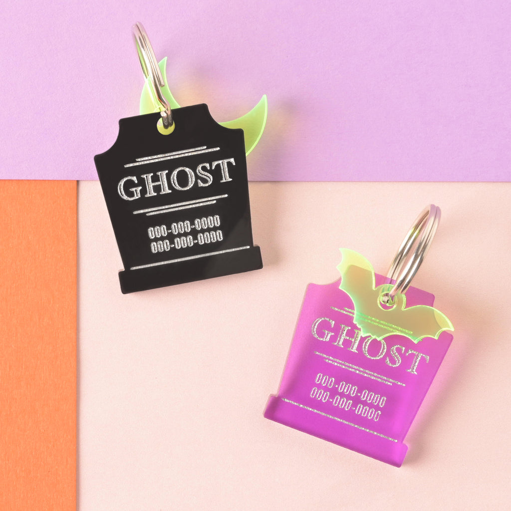 A product photo showing two custom-engraved tombstone-shaped pet ID tags on a colorful, split background (purple, pink, and orange). The tag on the top is black, engraved with "GHOST" in white, and features a neon yellow/green crescent moon charm. The tag on the bottom is purple, engraved with "GHOST" in a shiny pink color, and features a neon yellow/green bat charm. Both tags are shown with a silver split key ring.