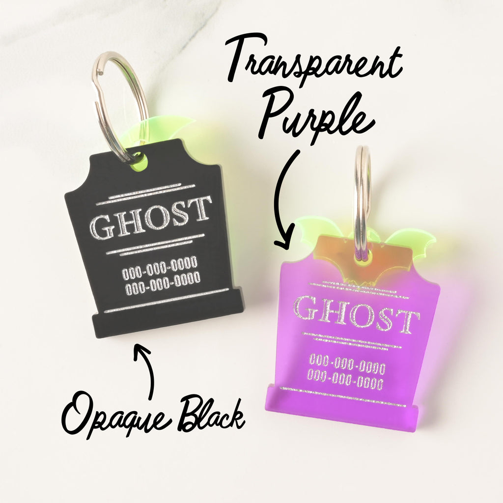 A product photo showing two custom tombstone-shaped pet ID tags. The tag on the left is Opaque Black, engraved with "GHOST" in white. The tag on the right is Transparent Purple, engraved with a shiny silver "GHOST." Both tags feature a neon yellow/green bat charm on top and a silver split key ring. Text labels indicate "Opaque Black" and "Transparent Purple."