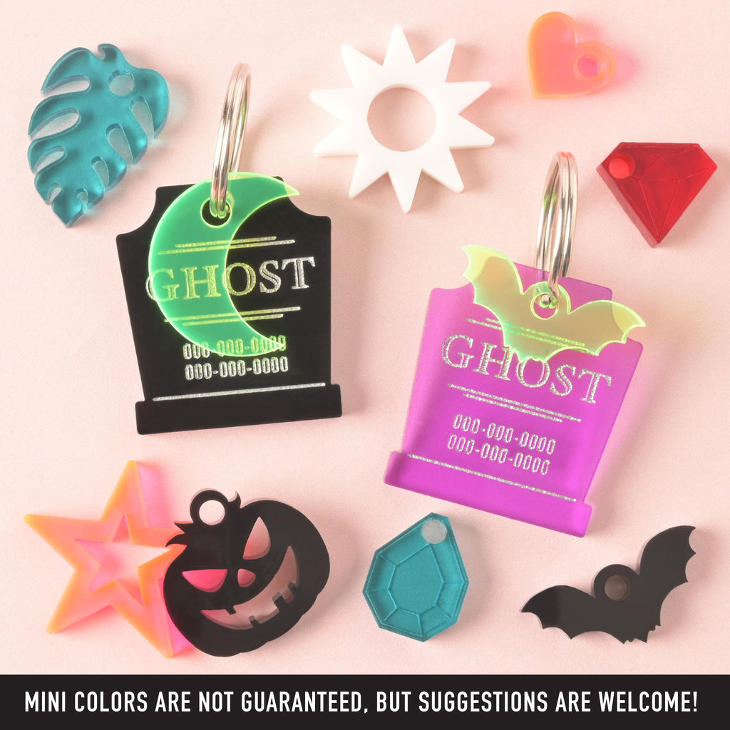 A product photo featuring two tombstone-shaped pet ID tags engraved with the word "GHOST." The tag on the left is black with a green crescent moon charm. The tag on the right is purple with a neon yellow/green bat charm. Surrounding the tags are various small charms: a teal monstera leaf, a white starburst sun, a neon orange heart, a red diamond gem, a black bat, a light blue geometric gem, a black jack-o'-lantern, and a neon orange star. Text at the bottom of the image reads, "MINI COLORS ARE NOT GUARANTEE