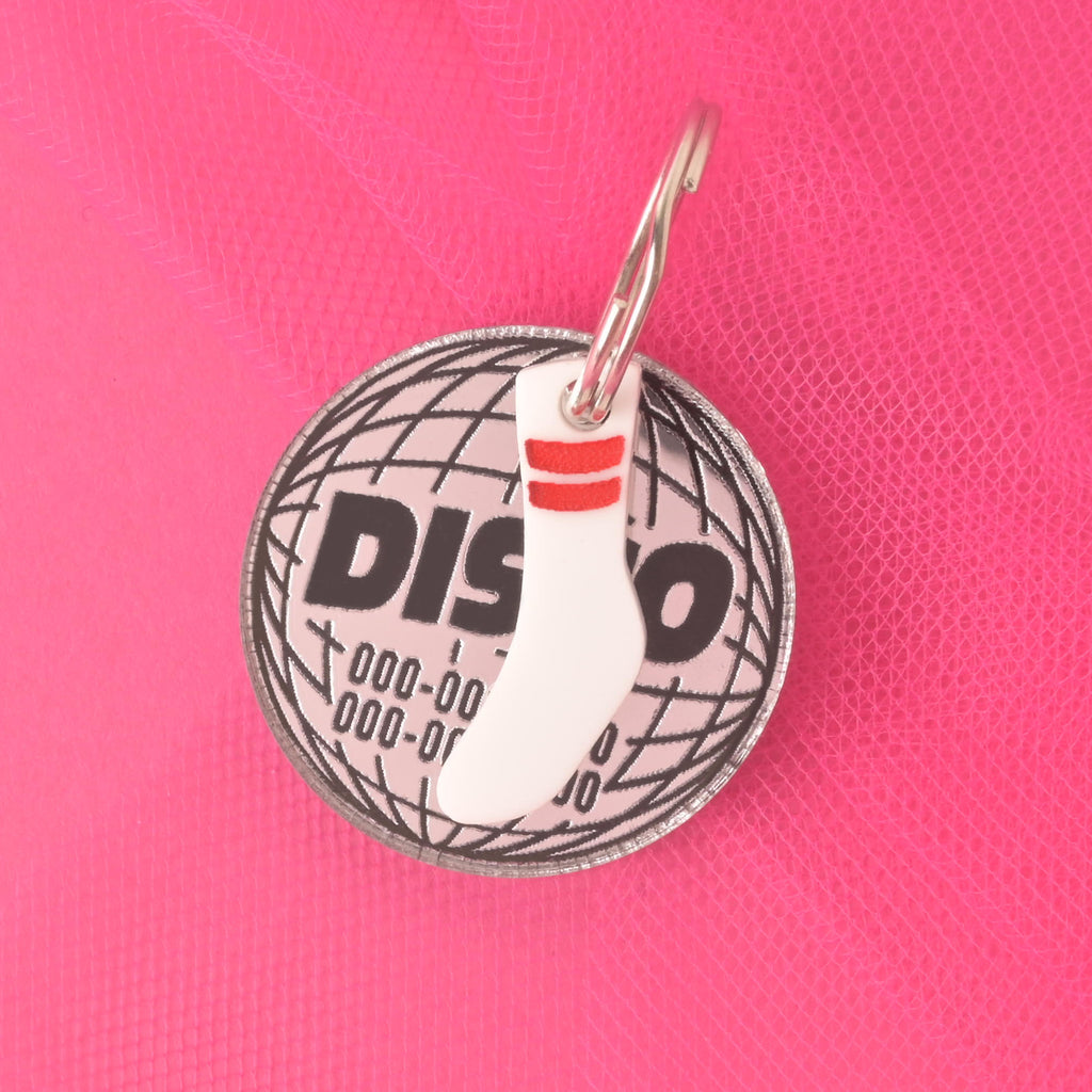A silver circular disco ball-shaped pet tag with the word "DISCO" and a placeholder phone number. A small white tube sock charm with red stripes is attached to the tag. Both are secured with a silver ring and are resting on a bright pink textured background.