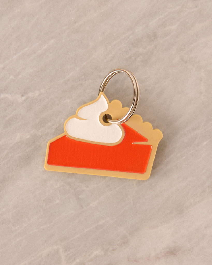 A novelty charm shaped like a slice of pie (pumpkin or sweet potato) made from layered acrylic in tan, red/orange, and white whipped cream. The charm is attached to a split ring and resting on a gray marble surface.