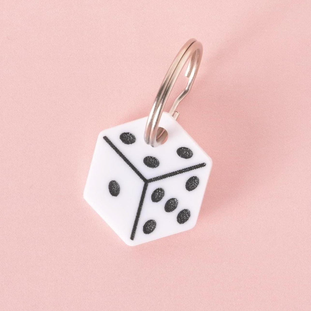 White dice-shaped pet ID tag with black dots and metal keyring, displayed on pink background.