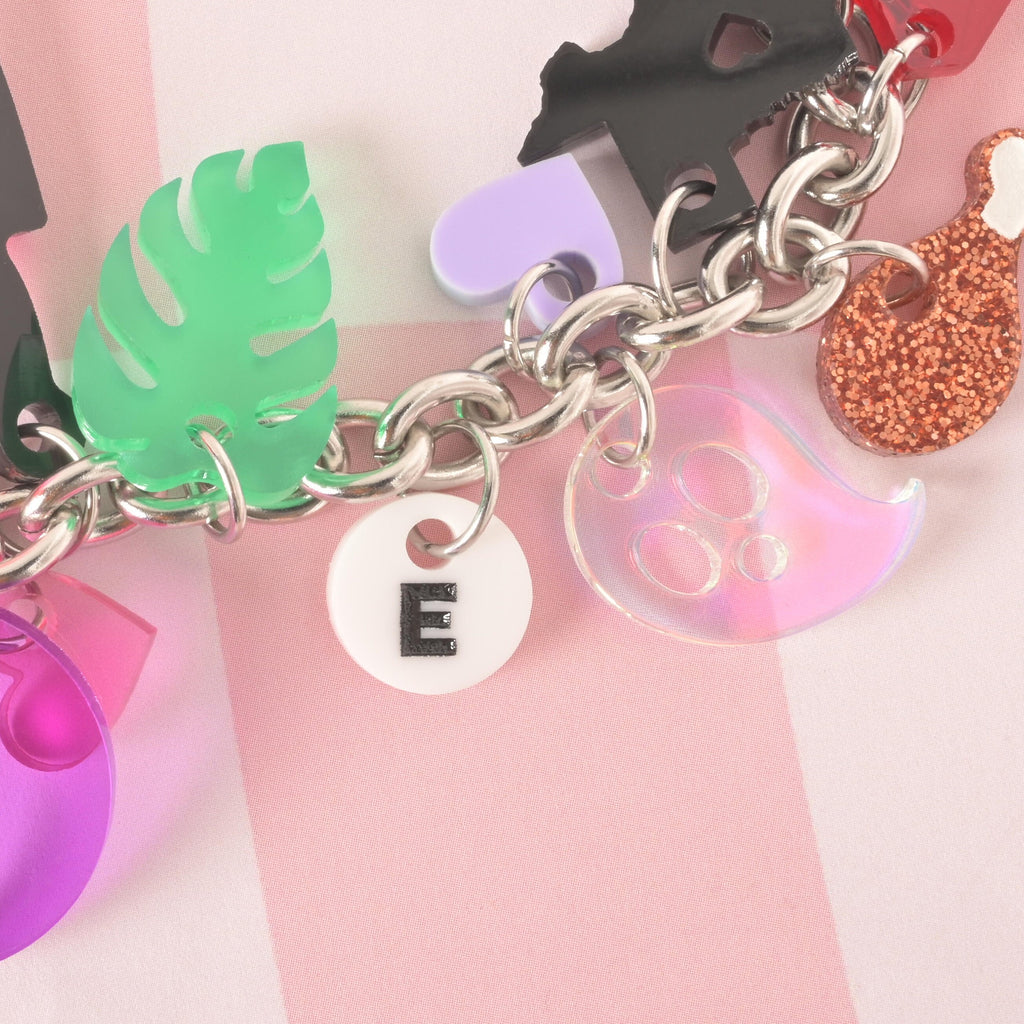 A close-up shot of a thick silver chain with several charms attached. The charms include a white circle with a black letter "E," a translucent iridescent ghost, a light green monstera leaf, a pink bow, a purple heart, and a glittery gold dog bone. The charms and chain are displayed on a pink and white checkered background.