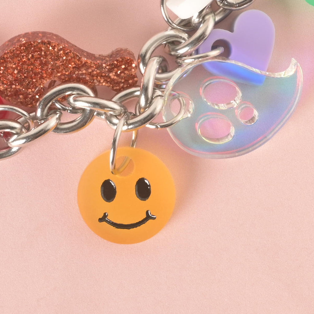A close-up shot of a thick silver chain with several charms attached. The charms include an orange smiley face, a translucent iridescent ghost, a purple-pink heart, and a glittery gold dog bone. The charms and chain are displayed on a light pink background.